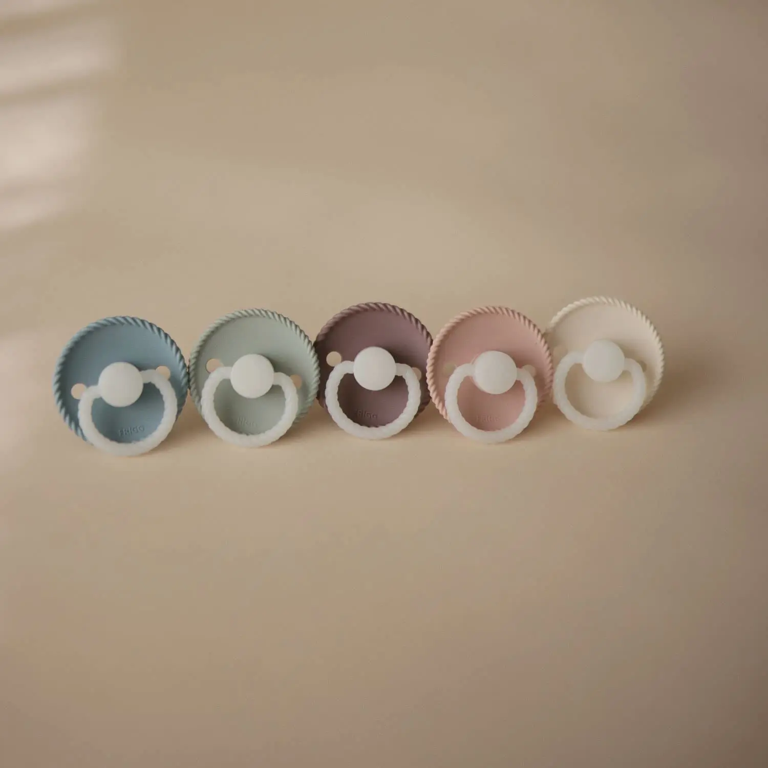 Two-pack of FRIGG Rope pacifiers in cream and stone blue with glow-in-the-dark handles and round, latex-free silicone nipples, designed to mimic breastfeeding.